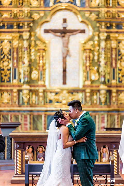Real-wedding-lani-and-ryan-kissing-in-the-church-his-hands-on-her-face-groom-in-green-suit-with-a-floral-tie-and-brown-shoes-bride-in-a-lace-a-line-dress-with-hair-in-a-bun-and-long-veil