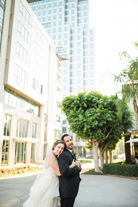 Avenue-of-the-arts-wedding-bride-hugging-groom-behind-him-bride-in-tulle-ball-gown-with-beaded-bodice-and-beaded-headband-groom-inblack-tuxedo-with-a-silver-vest-and-silver-bowtie