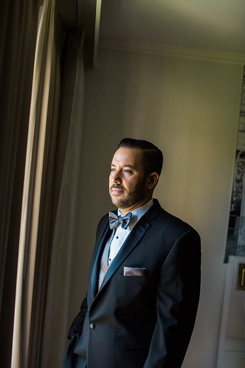 Avenue-of-the-arts-wedding-groom-looking-out-window-bride-in-tulle-ball-gown-with-beaded-bodice-and-beaded-headband-groom-inblack-tuxedo-with-a-silver-vest-and-silver-bowtie