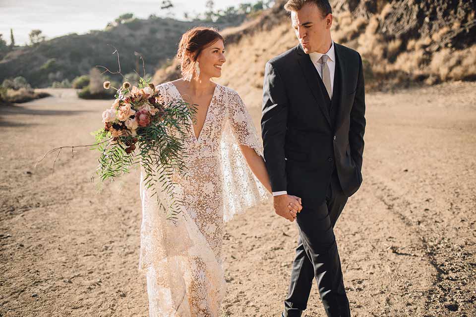 bronson-caves-elopement-shoot-bride-and-groom-walking-bride-wearing-a-lace-boho-inspired-dresswith-a-flowing-cape-detail-and-hair-up-in-a-mess-braided-bun-the-groom-wore-a-navy-suit-with-a-silver-tie