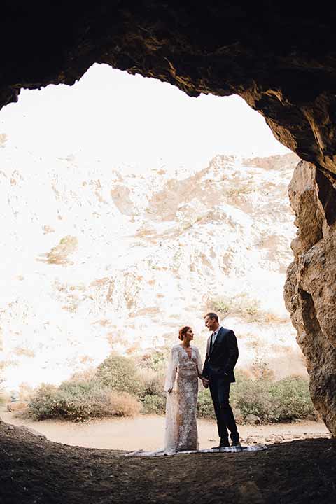 bronson-caves-elopement-shoot-bride-and-groom-in-cave-in-background-bride-wearing-a-lace-boho-inspired-dresswith-a-flowing-cape-detail-and-hair-up-in-a-mess-braided-bun-the-groom-wore-a-navy-suit-with-a-silver-tie