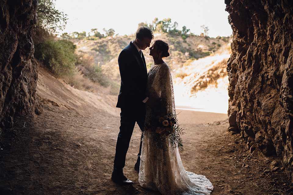 bronson-caves-elopement-shoot-bride-and-groom-in-cave-bride-wearing-a-lace-boho-inspired-dresswith-a-flowing-cape-detail-and-hair-up-in-a-mess-braided-bun-the-groom-wore-a-navy-suit-with-a-silver-tie