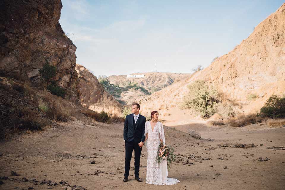 bronson-caves-elopement-shoot-bride-and-groom-looking-in-opposite-directions-bride-wearing-a-lace-boho-inspired-dresswith-a-flowing-cape-detail-and-hair-up-in-a-mess-braided-bun-the-groom-wore-a-navy-suit-with-a-silver-tie