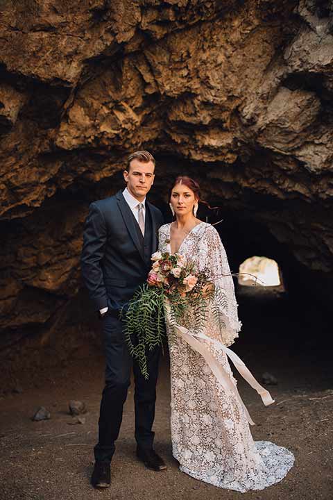 bronson-caves-elopement-shoot-bride-and-groom-outside-of-cave-looking-at-camera-bride-wearing-a-lace-boho-inspired-dresswith-a-flowing-cape-detail-and-hair-up-in-a-mess-braided-bun-the-groom-wore-a-navy-suit-with-a-silver-tie