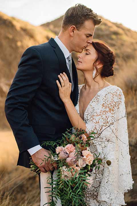 bronson-caves-elopement-shoot-bride-and-groom-with-sunset-behind-them-bride-wearing-a-lace-boho-inspired-dresswith-a-flowing-cape-detail-and-hair-up-in-a-mess-braided-bun-the-groom-wore-a-navy-suit-with-a-silver-tie