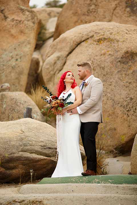 Desert-view-tower-bride-and-groom-embrace-on-rock-bride-in-a-fitted-satin-dress-with-a-high-neckline-and-hot-pink-hair-groom-in-a-tan-coat-with-black-pants-and-a-blue-tie