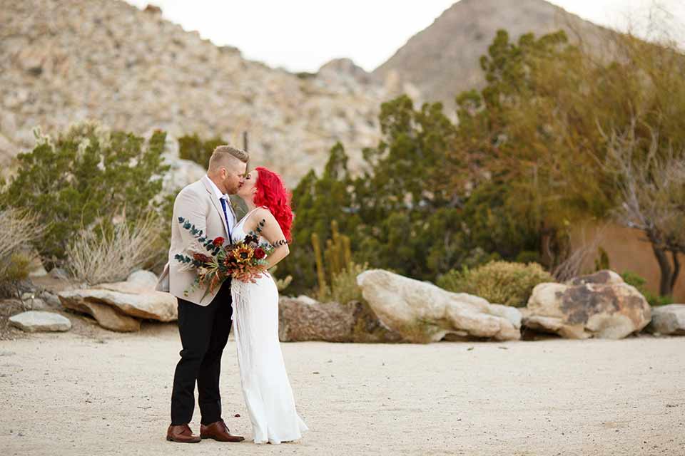 Desert-view-tower-bride-and-groom-kissing-bride-in-a-fitted-satin-dress-with-a-high-neckline-and-hot-pink-hair-groom-in-a-tan-coat-with-black-pants-and-a-blue-tie