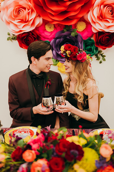 dia-de-los-muertos-cheers-bride-in-a-black-dress-with-floral-headpiece-groom-in-burgundy-tux-with-black-shirt-and-bow-tie