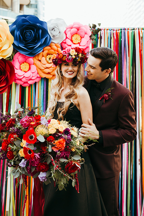 dia-de-los-muertos-color-background-bride-wearing-a-black-dress-and-floral-headpeice-groom-wearing-a-burgundy-tux-with-black-tri-and-black-shirt-with-black-bow-tie