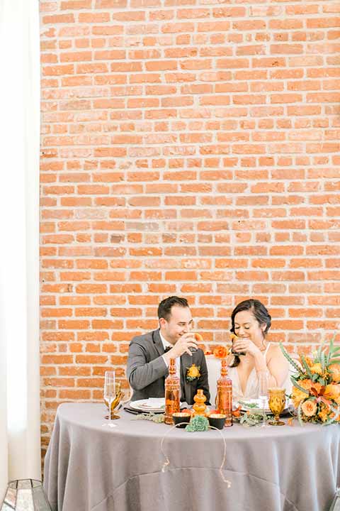 estate-on-second-shoot-bride-and-groom-at-sweetheart-table-bride-wearing-a-flowing-white-gown-with-thin-straps-and-hair-in-a-messy-boho-french-braid-groom-in-a-charcoal-grey-tuxedo-with-a-dark-brown-bow-and-shoes