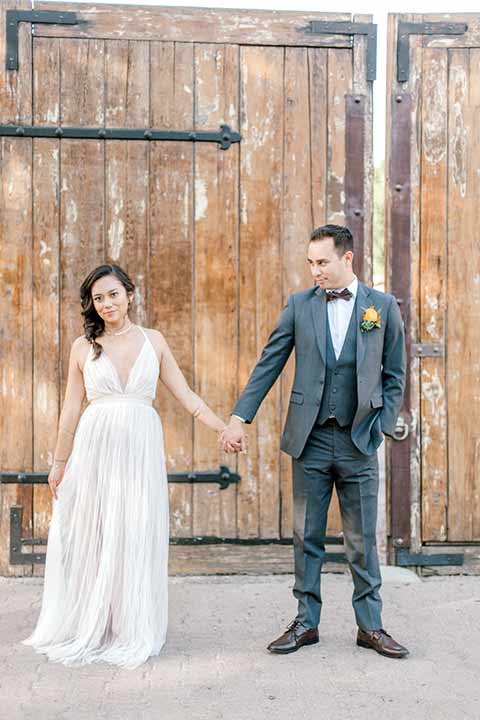 estate-on-second-shoot-bride-and-groom-holding-hands-bride-wearing-a-flowing-white-gown-with-thin-straps-and-hair-in-a-messy-boho-french-braid-groom-in-a-charcoal-grey-tuxedo-with-a-dark-brown-bow-and-shoes