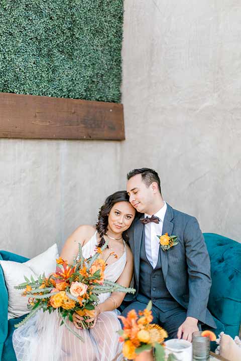 estate-on-second-shoot-bride-and-groom-on-blue-sofa-bride-looking-at-camera-bride-wearing-a-flowing-white-gown-with-thin-straps-and-hair-in-a-messy-boho-french-braid-groom-in-a-charcoal-grey-tuxedo-with-a-dark-brown-bow-and-shoes