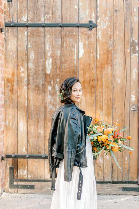 estate-on-second-shoot-bride-with-leather-jacket-bride-wearing-a-flowing-white-gown-with-thin-straps-and-hair-in-a-messy-boho-french-braid 