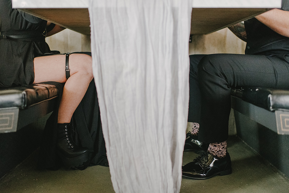 kindred-venue-gothic-inspired-shoot-bride-and-groom-legs-under-table-bride-in-black-3/4-sleeve-dress-with-strap-and-metal-detailing-with-a-black-wide-brimmed-hat-groom-in-all-black-tuxedo-with-black-accessories