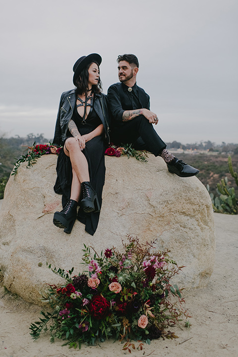 kindred-venue-gothic-inspired-shoot-bride-and-groom-on-rock-bride-in-black-3/4-sleeve-dress-with-strap-and-metal-detailing-with-a-black-wide-brimmed-hat-groom-in-all-black-tuxedo-with-black-accessories