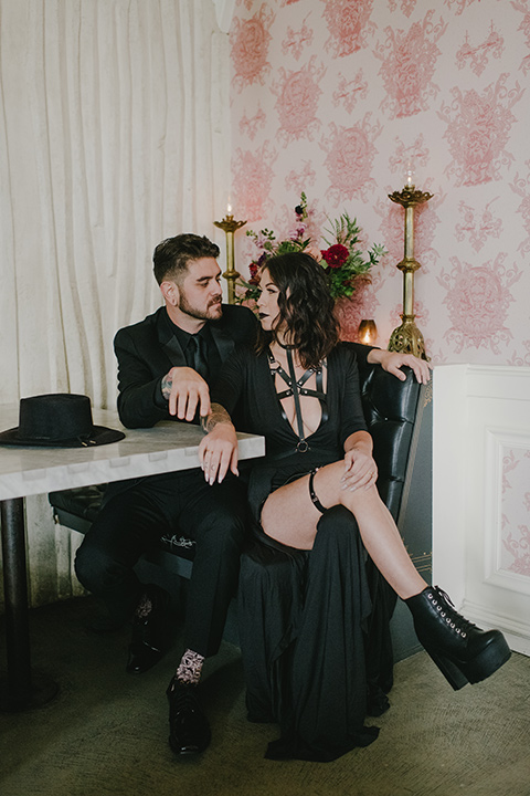 kindred-venue-gothic-inspired-shoot-bride-and-groom-sitting-bride-legs-crossed-bride-in-black-3/4-sleeve-dress-with-strap-and-metal-detailing-with-a-black-wide-brimmed-hat-groom-in-all-black-tuxedo-with-black-accessories