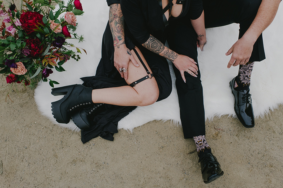 kindred-venue-gothic-inspired-shoot-close-up-of-legs-on-fuzzy-blanket-bride-in-black-3/4-sleeve-dress-with-strap-and-metal-detailing-with-a-black-wide-brimmed-hat-groom-in-all-black-tuxedo-with-black-accessories