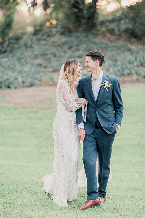 MUCKENTHALER-SHOOT-bride-and-groom-walking-connected-hand-in-hand-bride-in-fitted-satin-long-sleeve-gown-groom-in-a-slate-blue-suit-with-a-ivory-bow-tie