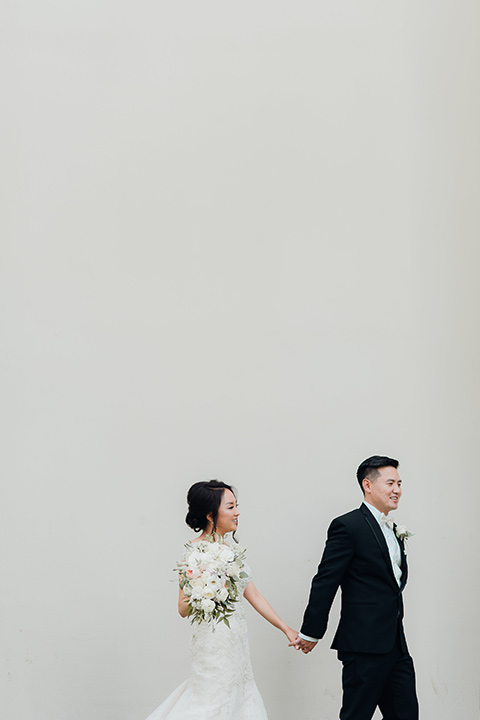 occidental-college-wedding-bride-and-groom-walking-by-blank-wal-groom-in-black-tuxedo-bride-in-a-fitted-gown-with-a-beaded-bodice