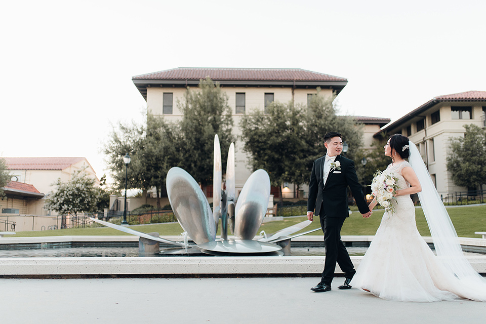 occidental-college-wedding-bride-and-groom-walking-by-fountain-groom-in-black-tuxedo-bride-in-a-fitted-gown-with-a-beaded-bodice