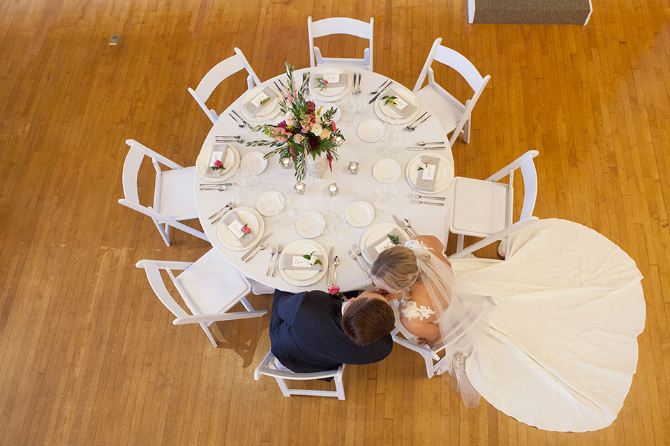 parq-west-shoot-couple-sitting-at-table -bride-wearing-a-flowing-gown-with-an-illusion-lace-bodice-and-cap-sleeves-groom-in-a-charcoal-tuxedo-with-a-black-satin-shawl-lapel