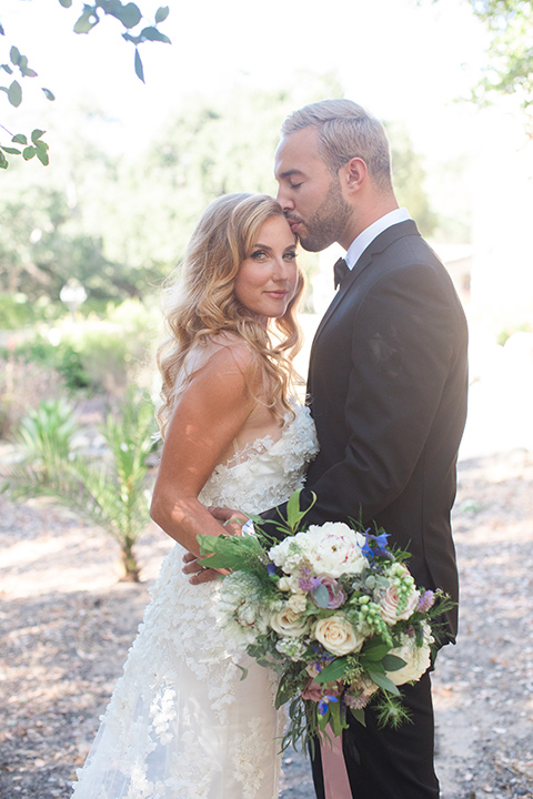 rockwood-shoot-bride-looking-at-camera-groom-kissing-her-head-outside-bride-in-a-ball-gown-with-hair-in-a-retro-inspired-finger-wave-groom-in-a-traditional-black-tuxedo-with-black-bow-tie