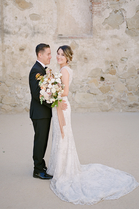 san-juan-capastrano-wedding-bride-looking-at-camera-groom-looking-at-her-bride-in-a-white-fitted-silk-dress-with-cap-sleeves-and-an-illusion-neckline-and-back-groom-in-a-black-tuxedo-with-black-bow-tie