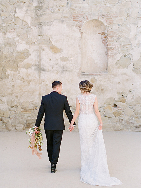 san-juan-capastrano-wedding-couple-walking-away-bride-in-a-white-fitted-silk-dress-with-cap-sleeves-and-an-illusion-neckline-and-back-groom-in-a-black-tuxedo-with-black-bow-tie