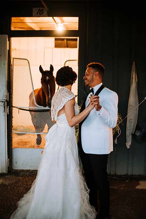 santa-anita-park-art-deco-shoot-bride-and-groom-with-horses-bride-wearing-a-vintage-inspired-dress-with-delicate-beading-and-flutter-sleeves-groom-wearing-a-white-tuxedo-jacket-with-black-trim-and-black-pants