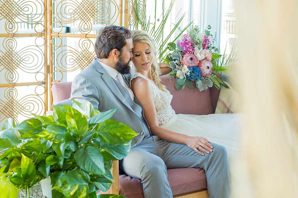 resized-seapoint-bridal-shoot-bride-and-groom-on-couch-bride-in-a-lace-gown-with-thick-lace-straps-and-hair-in-a-loose-braid-groom-in-a-light-grey-suit-with-a-white-long-tie