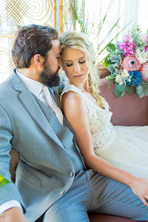 seapoint-bridal-shoot-bride-and-groom-sitting-eyes-closed-bride-in-a-lace-gown-with-thick-lace-straps-and-hair-in-a-loose-braid-groom-in-a-light-grey-suit-with-a-white-long-tie