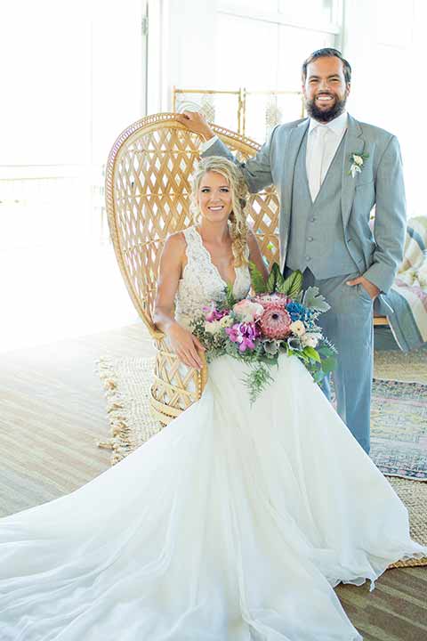 seapoint-bridal-shoot-bride-in-chair-groom-next-to-her-bride-in-a-lace-gown-with-thick-lace-straps-and-hair-in-a-loose-braid-groom-in-a-light-grey-suit-with-a-white-long-tie