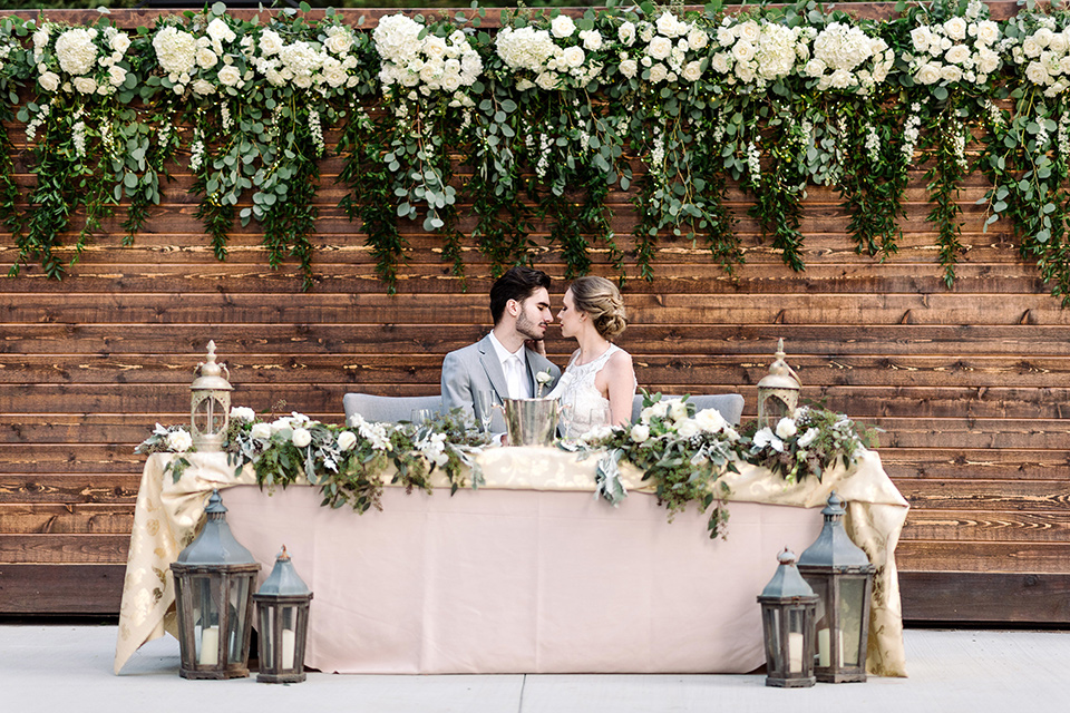 skypark-styled-shoot-bride-and-groom-sweetheart-table-bride-in-lace-fitted-dress-with-ca-sleeves-and-a-keyhole-back-detail-groom-in-grey-suit-with-brown-shoes-and-ivory-long-tie