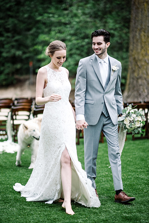 skypark-styled-shoot-bride-and-groom-walking-dog-behind-them-bride-in-lace-fitted-dress-with-a-sleeves-and-a-keyhole-back-detail-groom-in-grey-suit-with-brown-shoes-and-ivory-long-tie