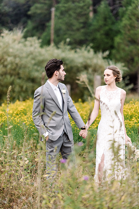 skypark-styled-shoot-bride-and-groom-walking-in-floral-fields-bride-in-lace-fitted-dress-with-a-sleeves-and-a-keyhole-back-detail-groom-in-grey-suit-with-brown-shoes-and-ivory-long-tie