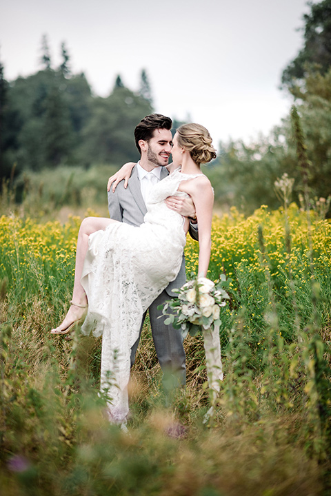 skypark-styled-shoot-groom-holding-bride-in-flowers-bride-in-lace-fitted-dress-with-a-sleeves-and-a-keyhole-back-detail-groom-in-grey-suit-with-brown-shoes-and-ivory-long-tie