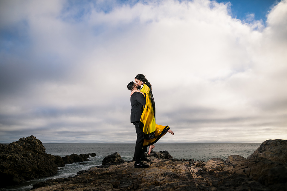 Terranea-styled-shoot-groom-holding-up-bride-kissing-bride-in-yellow-bride-in-yellow-sarees-with-hair-in-a-loose-wave-groom-in-a-tradtitional-black-tuxedo-with-a-black-bowtie-and-black-vest-with-wearing-a-black-and-yellow-florals