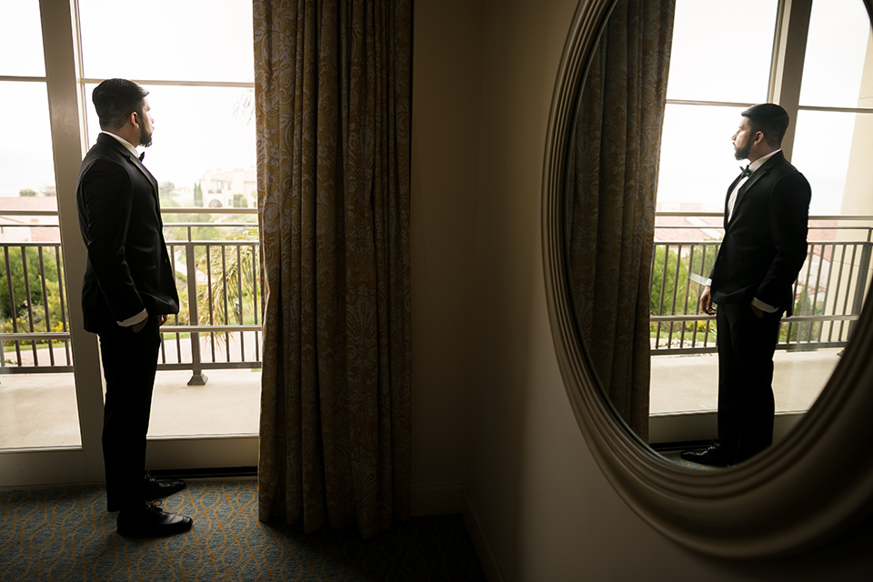 Terranea-styled-shoot-groom-looking-out-the-window-with-mirror-photo-groom-in-a-tradtitional-black-tuxedo-with-a-black-bowtie-and-black-vest