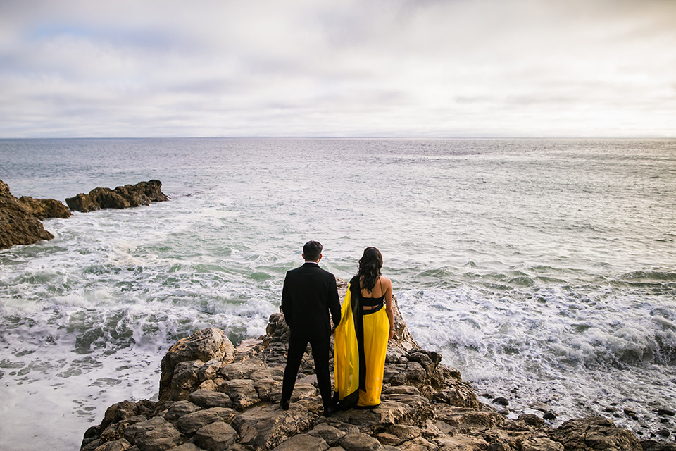 Terranea-styled-shoot-on-the-water-both-facing-the-ocean-bride-kissing-bride-in-yellow-bride-in-yellow-sarees-with-hair-in-a-loose-wave-groom-in-a-tradtitional-black-tuxedo-with-a-black-bowtie-and-black-vest-with-wearing-a-black-and-yellow-florals