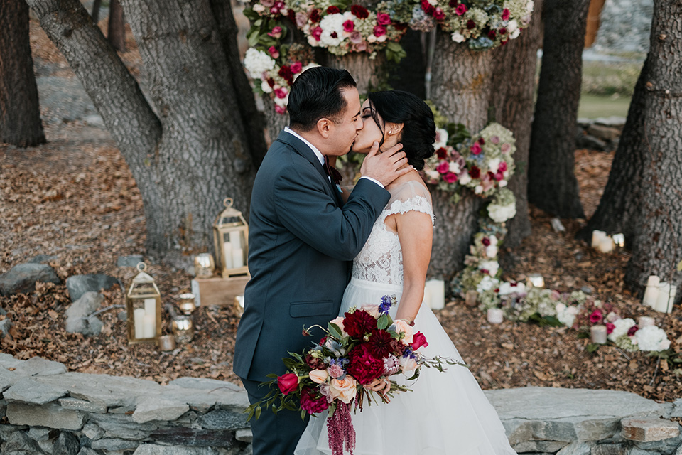the-guest-ranch-ceremony-kiss-groom-wearing-a-dark-grey-suit-with-light-grey-vest-and-a-burgundy-bowtie-bride-in-a-tulle-ballgown-with-straps-and-a-beaded-bodice