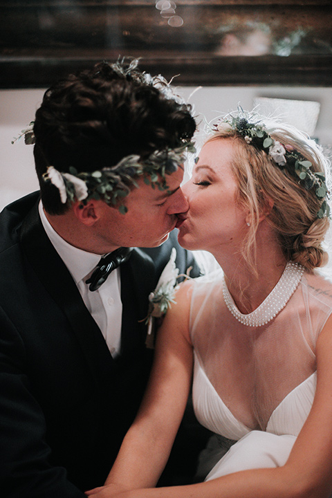 Voila-studios-bride-and-groom-kissing-with-floral-crowns-bride-in-a-flowing-gown-with-a-natural-waistline-and-an-illusion-neckline-with-a-floral-crown-groom-in-a-navy-tuxedo-with-a-leather-bow-tie