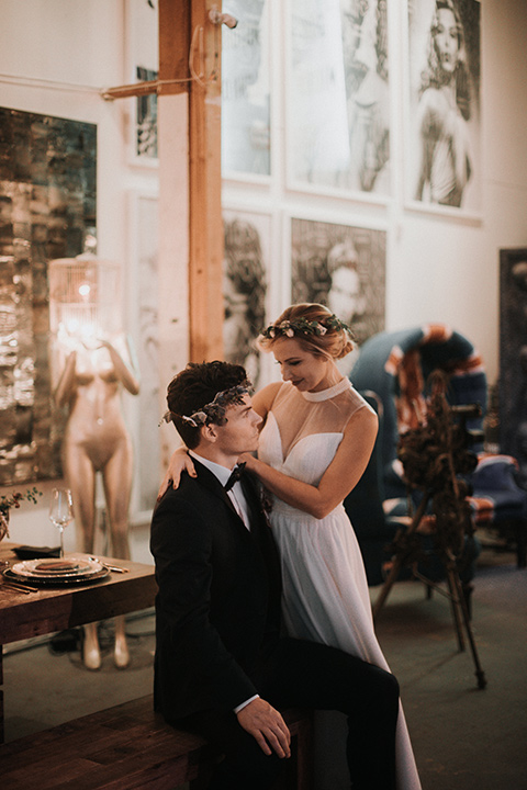 Voila-studios-bride-standing-next-to-groom-sitting-bride-in-a-flowing-gown-with-a-natural-waistline-and-an-illusion-neckline-with-a-floral-crown-groom-in-a-navy-tuxedo-with-a-leather-bow-tie