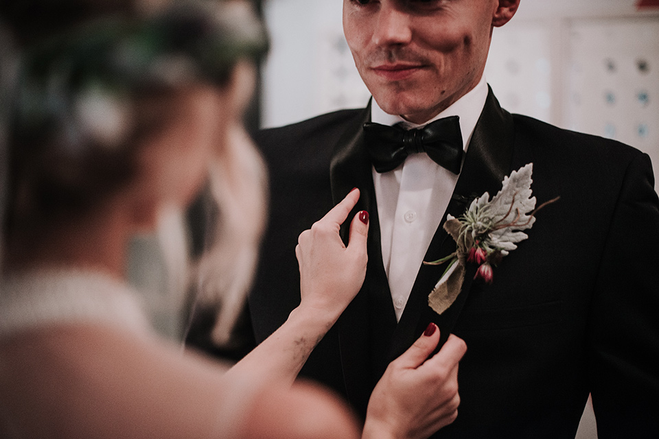 Voila-studios-bride-touching-jacket-bride-in-a-flowing-gown-with-a-natural-waistline-and-illusion-neckline-and-floral-crown-groom-in-a-navy-tuxedo-with-leather-bow-tie