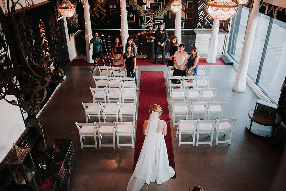 Voila-studios-bride-waling-down-the-aisle-bride-in-a-flowing-gown-with-a-natural-waistline-and-illusion-neckline-and-floral-crown-groom-in-a-navy-tuxedo-with-leather-bow-tie