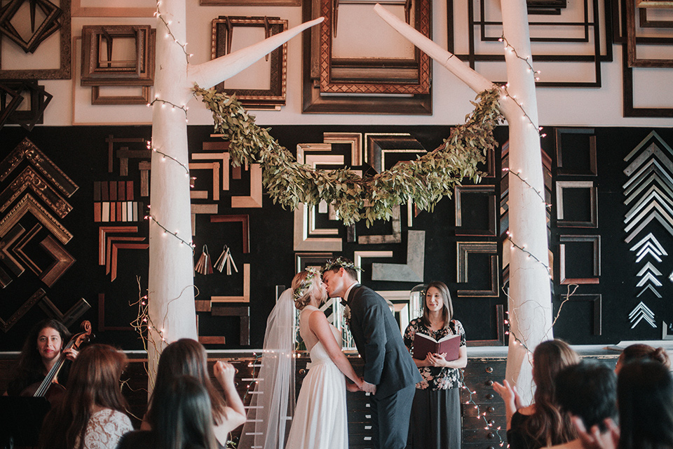 Voila-studios-ceremony-kiss-bride-in-a-flowing-gown-with-a-natural-waistline-and-illusion-neckline-and-floral-crown-groom-in-a-navy-tuxedo-with-leather-bow-tie