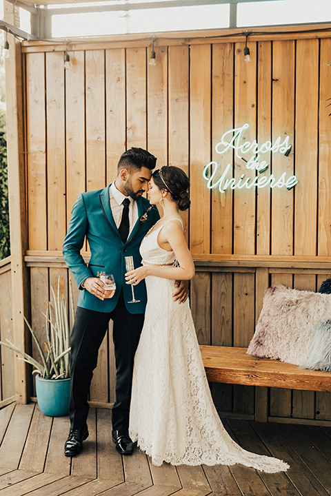 The-Ruby-Street-photoshoot-bride-and-groom-by-wooden-wall-groom-in-a-teal-tuxedo-jacket-with-black-pants-and-a-black-bowtie-bride-in-a-lace-dress-with-a-deep-v-neckline