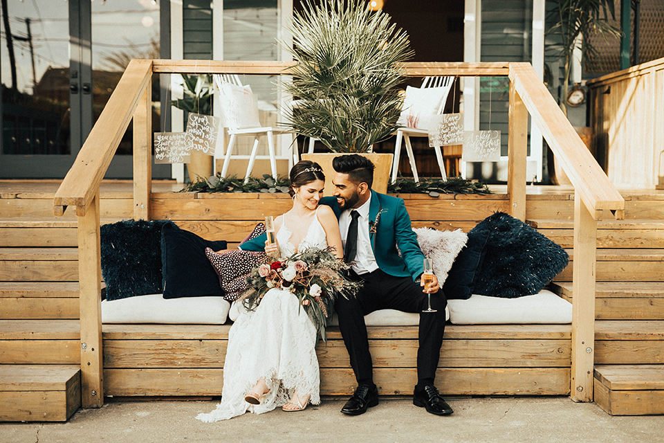 The-Ruby-Street-photoshoot-bride-and-groom-sitting-outside-groom-in-a-teal-tuxedo-with-black-pants-and-bride-in-a-lace-bohemian-dress