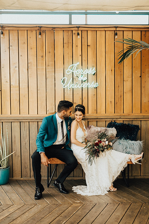 The-Ruby-Street-photoshoot-bride-and-groom-sitting-under-neon-lights-groom-wearing-a-teal-jacket-with-black-pants-bride-wearing-a-lace-bohemian-style-dress