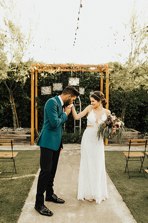 The-Ruby-Street-photoshoot-groom-kissing-brides-hand-groom-wearing-a-teal-jacket-with-black-pants-bride-wearing-a-lace-bohemian-style-dress