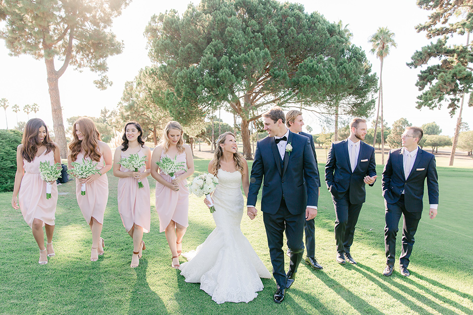 Los-Verdes-Golf-Course-Wedding-bridalparty-walking-bridesmaids-in-light-pink-short-dresses-groomsmen-in-blue-suits-with-light-pink-bowties-bride-is-in-a-lace-mermaid-style-gown-with-a-modified-sweetheart-neckline-the-groom-is-in-a-navy-blue-tuxedo-with-a-black-shawl-lapel-and-a-black-bow-tie